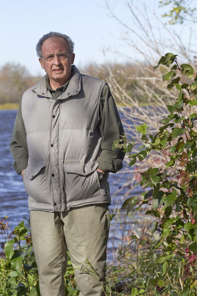 A man stands in front of a a lake next with his hands in his pockets facing the camera