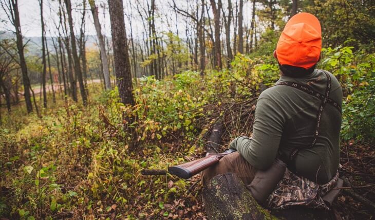 Person wearing orange cap and holding a rifle across their lap sits in a wooded area facing away from the camera
