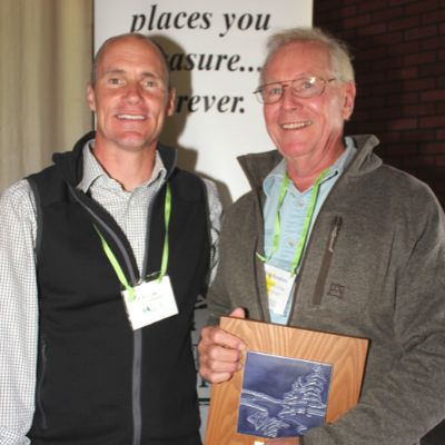 Two men pose for a picture, smiling for the camera, the second holds an award plaque