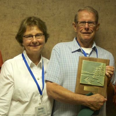 A married couple poses for a picture, smiling for the camera and the gentleman holds an award plaque