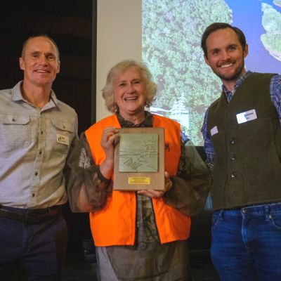 Three people pose for a picture together, smiling, the woman in the middle wears an orange vest and holds an award plaque