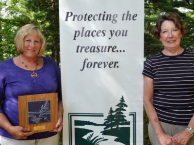 Two women smiling into the camera stand next to a sign reading, "Protecting the places you treasure...forever," one holding an award plaque