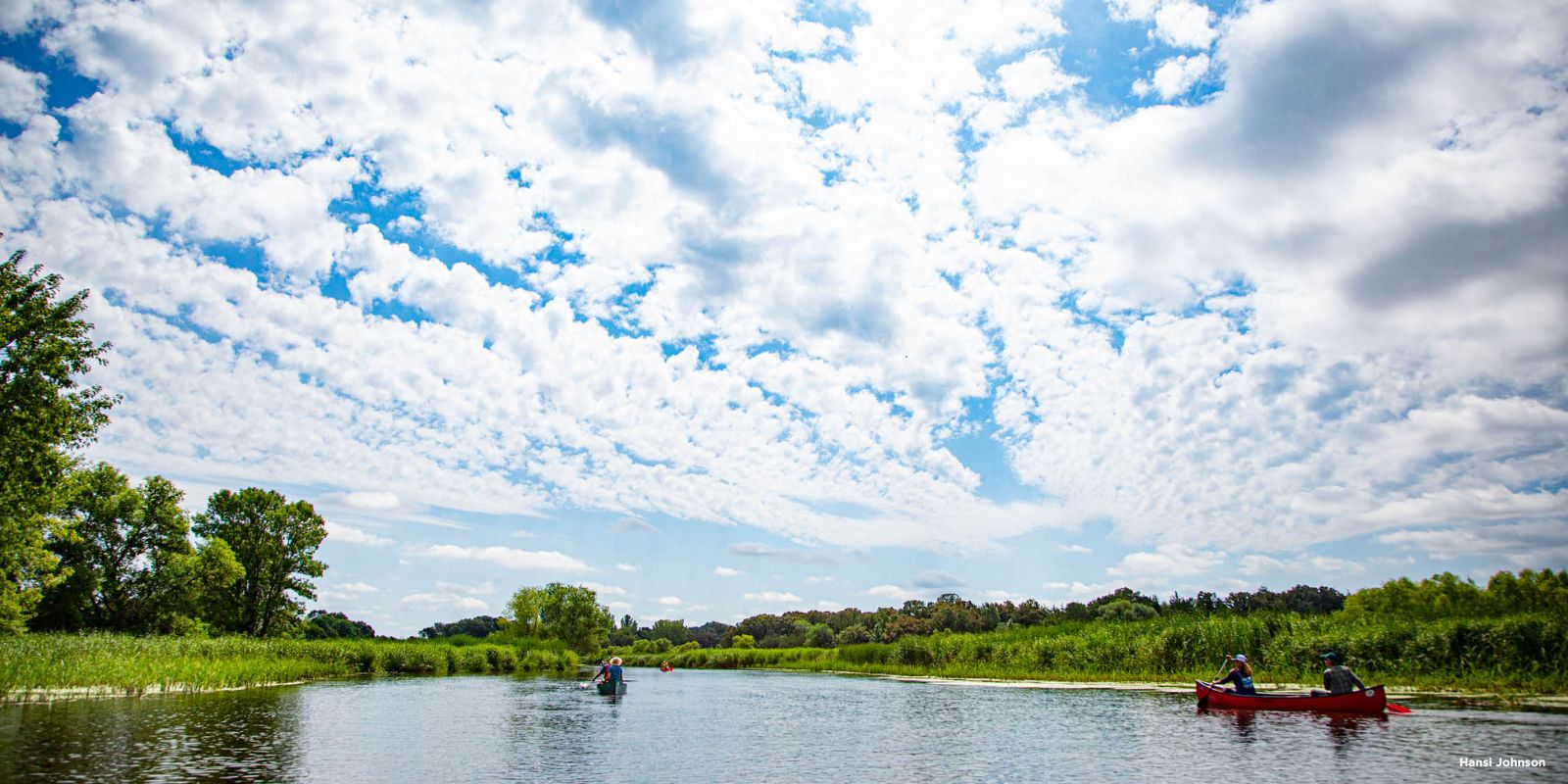 People paddle in canoes on the Sauk River with a verdant green shoreline on either side and blue sky