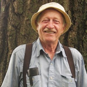 A man wearing a hat standing in front of a large tree smiles into the camera