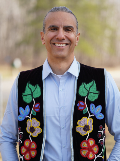An Ojibwe man wearing a traditional floral patterned vest smiles into the camera